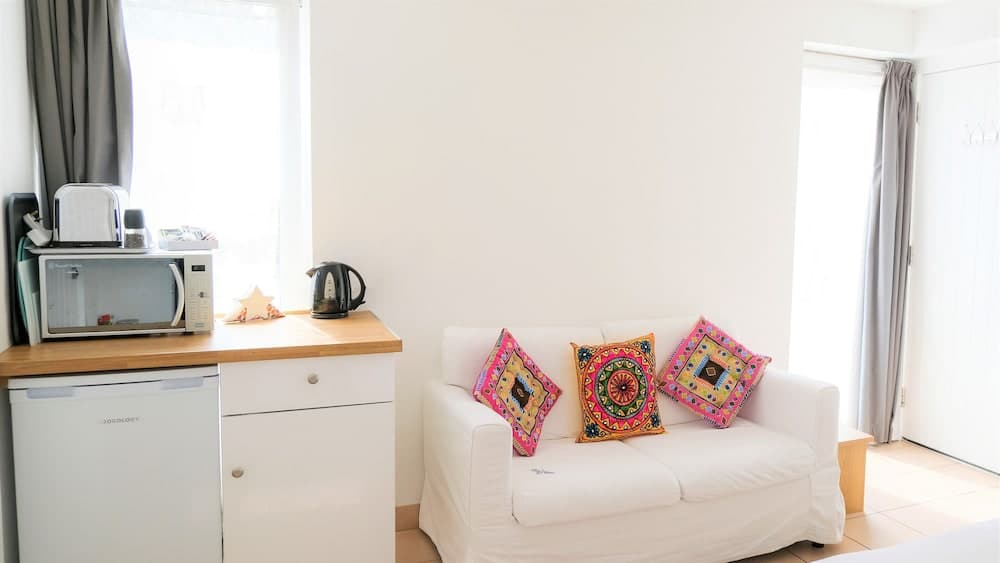 A cozy apartment living space features a white sofa adorned with vibrant patterned cushions, a compact kitchenette with a microwave and kettle, and soft natural light filtering through sheer curtains, ideal for travelers seeking a comfortable and stylish short-term rental.
