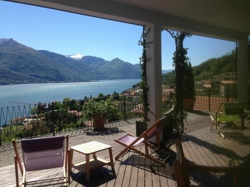 A serene lakeside terrace with wooden chairs and lush greenery offers breathtaking views of a tranquil lake surrounded by majestic mountains, ideal for a relaxing getaway or vacation rental listing.