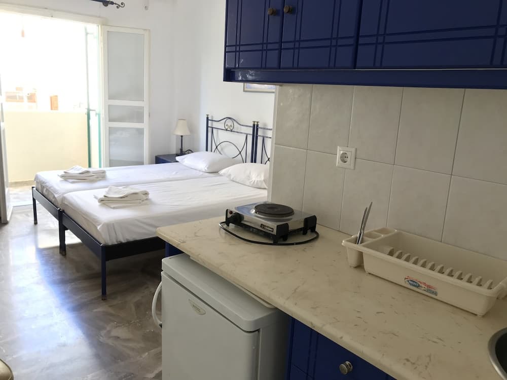 A cozy studio apartment features a compact kitchenette with blue cabinets and a countertop stove, adjacent to twin beds with white linens, ideal for budget-friendly travelers seeking comfort and convenience.