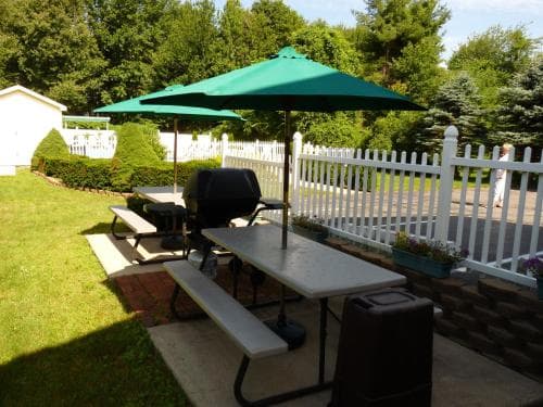 A serene backyard patio features picnic tables under green umbrellas, a white picket fence, lush greenery, and a barbecue grill, ideal for family gatherings or outdoor relaxation.
