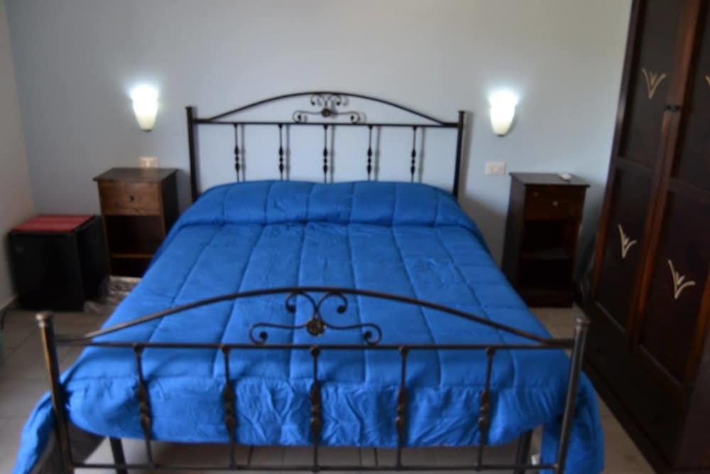 A cozy bedroom features a blue-covered bed with an ornate metal frame, flanked by wooden nightstands and wall-mounted lamps, ideal for travelers seeking a comfortable and simple accommodation.