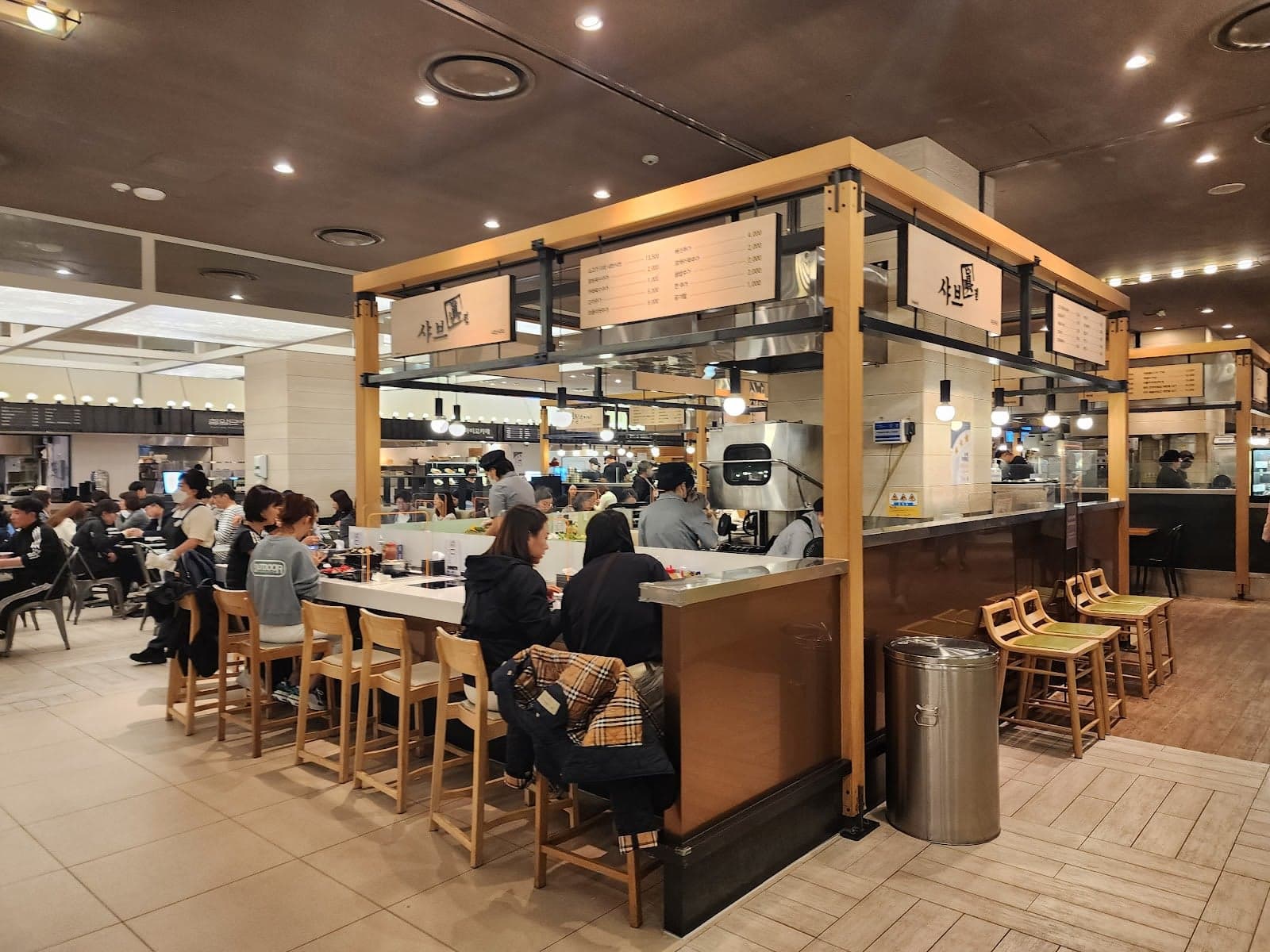 A bustling food court at Shinsegae Department Store in Busan, South Korea, features modern kiosks, wooden chairs, and a lively atmosphere perfect for travelers seeking local cuisine and shopping experiences.