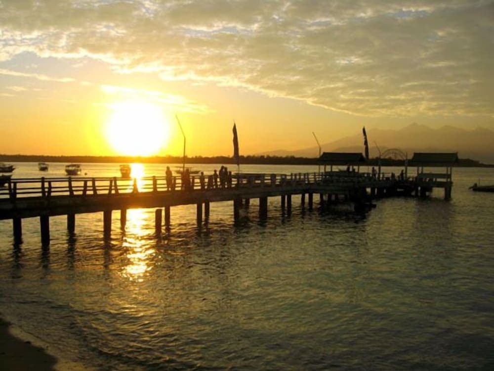A serene sunset over a tranquil waterfront features silhouetted figures on a wooden pier, distant boats, and mountains in the background, ideal for travelers seeking peaceful coastal retreats.