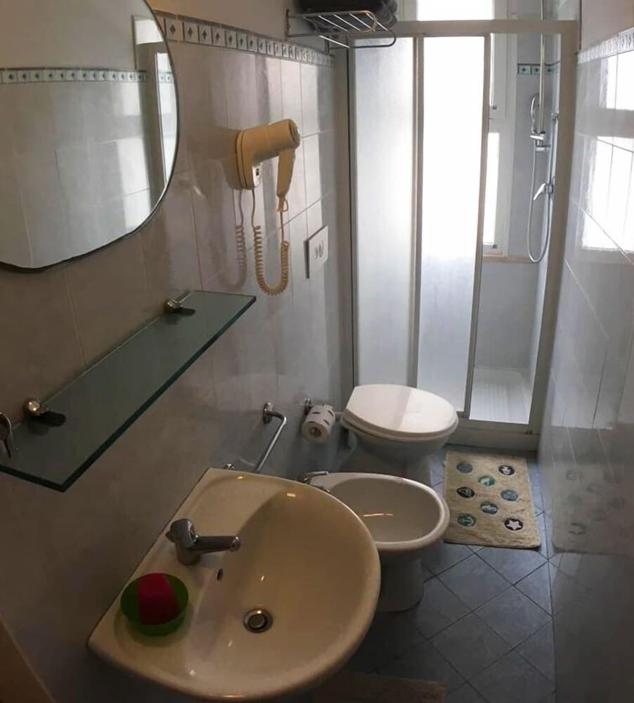 A cozy hotel bathroom features a sink with colorful cups, a wall-mounted hairdryer, a bidet, a toilet, and a glass-enclosed shower, creating a functional and inviting space for travelers seeking comfort.