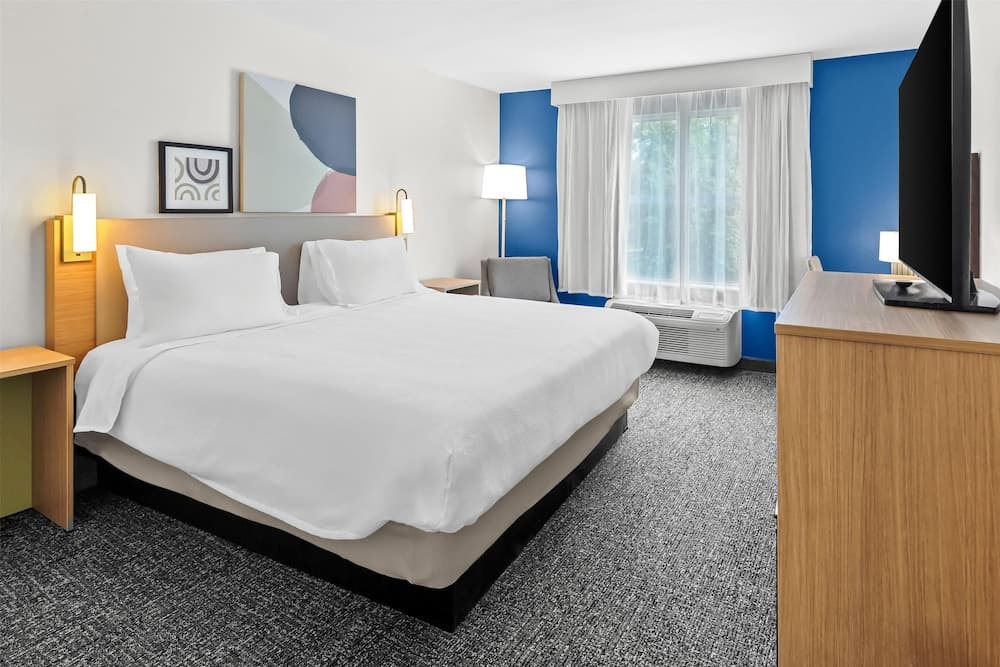 A cozy hotel room features a neatly made bed with white linens, modern artwork on the walls, a flat-screen TV on a wooden dresser, and a bright window with sheer curtains, ideal for travelers seeking comfort and relaxation.