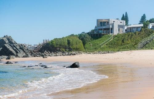 A serene coastal beach scene features gentle waves lapping onto sandy shores, modern houses perched on a hillside amidst lush greenery, and rocky outcrops, ideal for travelers seeking relaxation and scenic beauty.