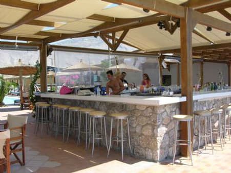 A serene outdoor bar with stone counters and metal stools under a wooden canopy invites travelers to relax by the poolside, perfect for a leisurely vacation vibe.