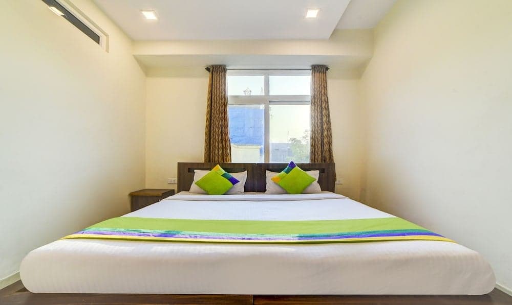 A cozy hotel room features a neatly made bed with vibrant green and purple accents, framed by patterned curtains and natural light from a large window, ideal for travelers seeking comfort and relaxation.