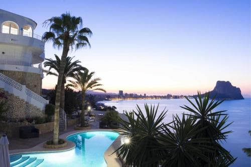 A serene coastal evening scene features a luxurious villa with a glowing pool, palm trees, and a stunning view of the sea and distant cliffs, ideal for a romantic getaway or luxury vacation.