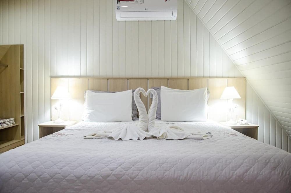 A cozy attic bedroom with white panel walls features a neatly made bed adorned with towel swans forming a heart, ideal for romantic getaways or honeymoon stays.