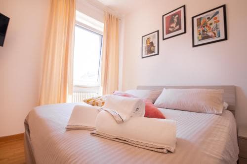 A cozy hotel room features a neatly made bed adorned with white linens and colorful pillows, framed by soft curtains and vibrant wall art, ideal for travelers seeking comfort and relaxation.