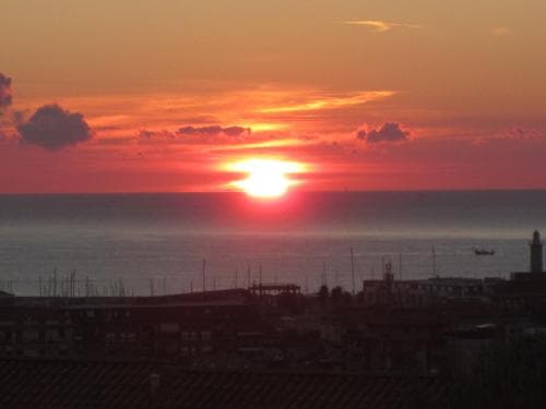 A serene sunset casts vibrant orange hues over a coastal cityscape, featuring silhouetted rooftops, a distant sailboat, and a tranquil sea, ideal for travelers seeking picturesque seaside views.