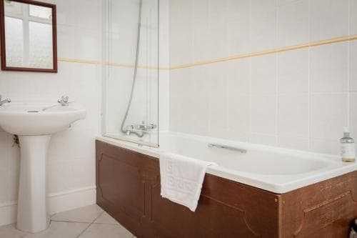 A cozy bathroom with white tiled walls, a wooden bathtub, a pedestal sink, and a mirror, ideal for travelers seeking a quaint and comfortable accommodation experience.