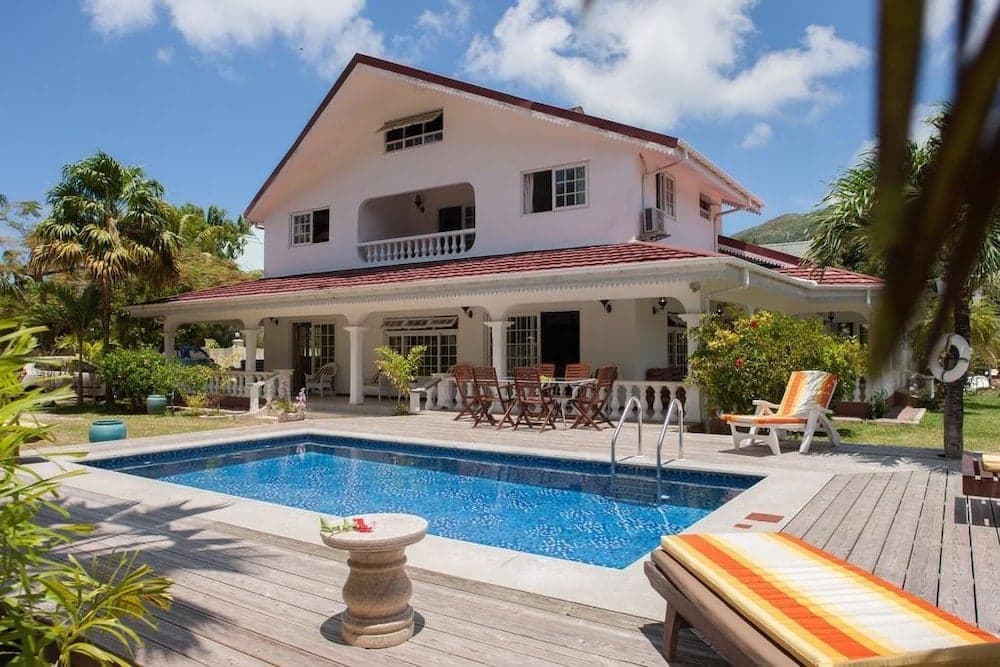 A serene tropical villa with a sparkling pool, wooden deck, lounge chairs, and lush greenery offers an idyllic vacation retreat under a bright blue sky.
