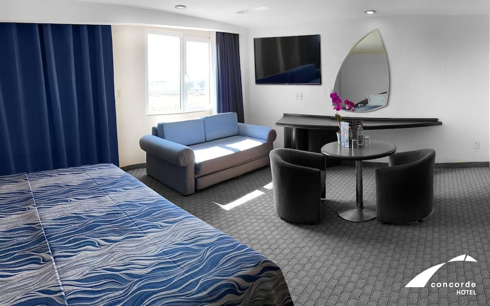 A cozy hotel room features a neatly made bed, a blue sofa by the window, a modern desk setup with a mirror, and a small dining area, ideal for travelers seeking comfort and functionality during their stay.