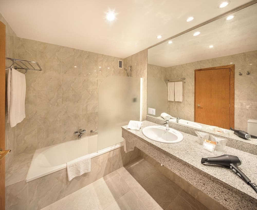 A serene hotel bathroom features a spacious bathtub, granite countertop with toiletries, large mirror, and hairdryer, ideal for travelers seeking comfort and convenience.