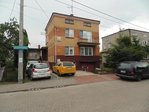 A quiet residential street lined with parked cars and modest apartment buildings under an overcast sky, ideal for travelers seeking affordable urban accommodations.