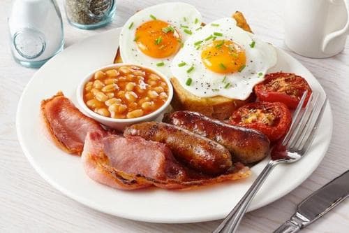 A hearty full English breakfast featuring sunny-side-up eggs, bacon, sausages, baked beans, grilled tomatoes, and hash browns on a white plate, perfect for a cozy morning meal at a quaint café or hotel dining room.