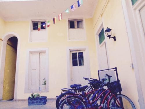 A charming courtyard with yellow walls, white doors, and windows adorned with colorful flags creates a cozy atmosphere, featuring parked bicycles and a potted plant, ideal for travelers seeking a quaint European hideaway.