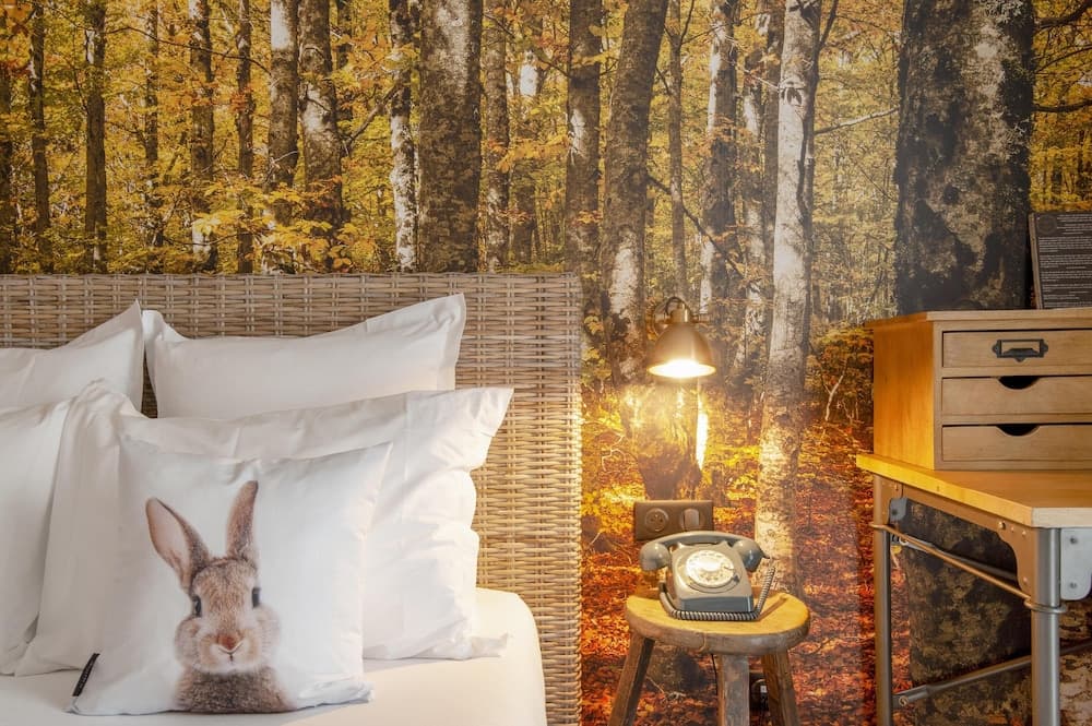 A cozy bedroom with a forest mural backdrop, featuring a wicker headboard, a rabbit pillow, a vintage telephone on a stool, and warm lighting, ideal for travelers seeking a serene nature-inspired retreat.