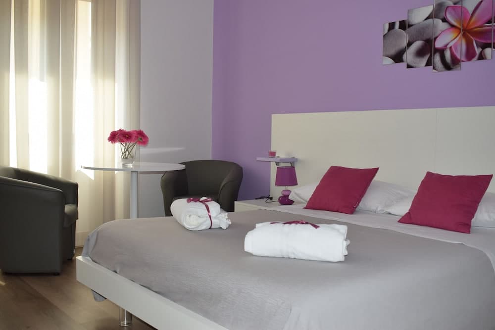 A cozy hotel room with purple walls, neatly arranged bed with white towels and pillows, a small table with flowers by the window, and modern decor, ideal for travelers seeking comfort and relaxation.