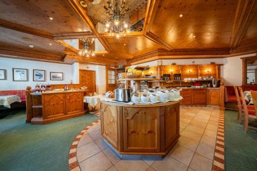 A cozy, warmly lit breakfast area with wooden furnishings, tiled floors, and a central serving counter invites travelers to enjoy a hearty start in a rustic lodge setting.