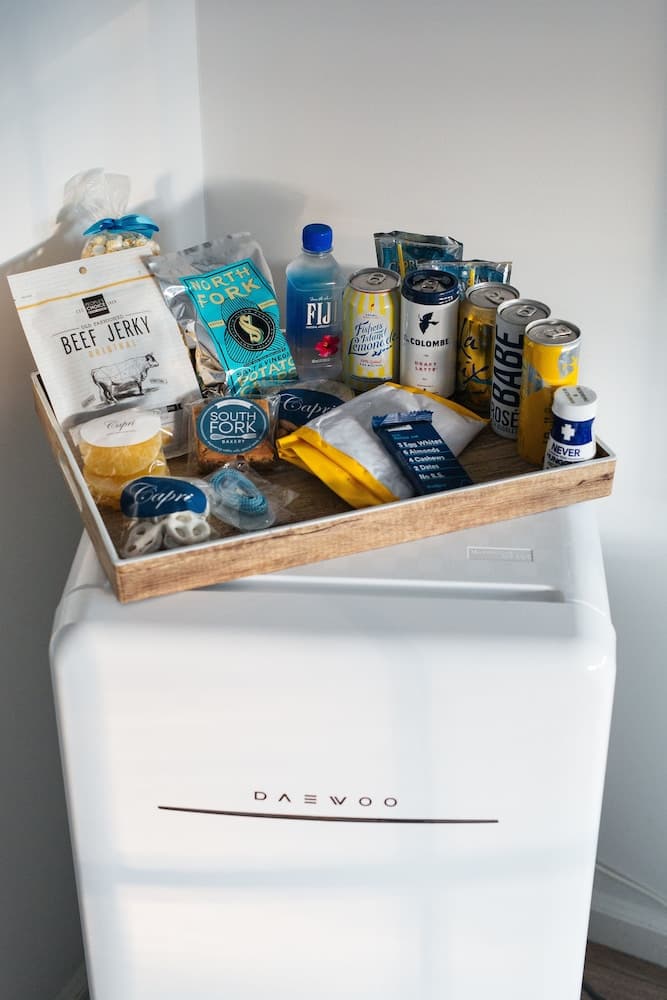 A cozy indoor setting features a wooden tray filled with snacks like beef jerky, potato chips, and beverages atop a sleek white Daewoo mini fridge, ideal for travelers seeking convenient refreshments in a compact space.