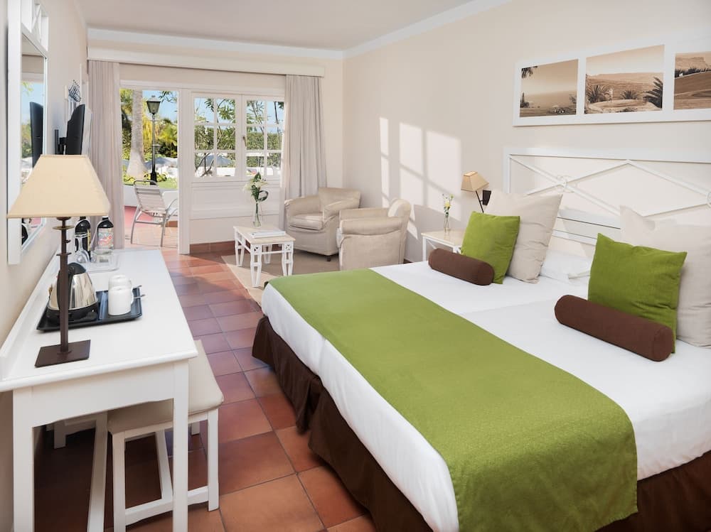 A cozy hotel room with a neatly made bed adorned with green and brown pillows, a desk with tea-making facilities, and a sunny balcony view of lush greenery, ideal for travelers seeking relaxation and comfort.