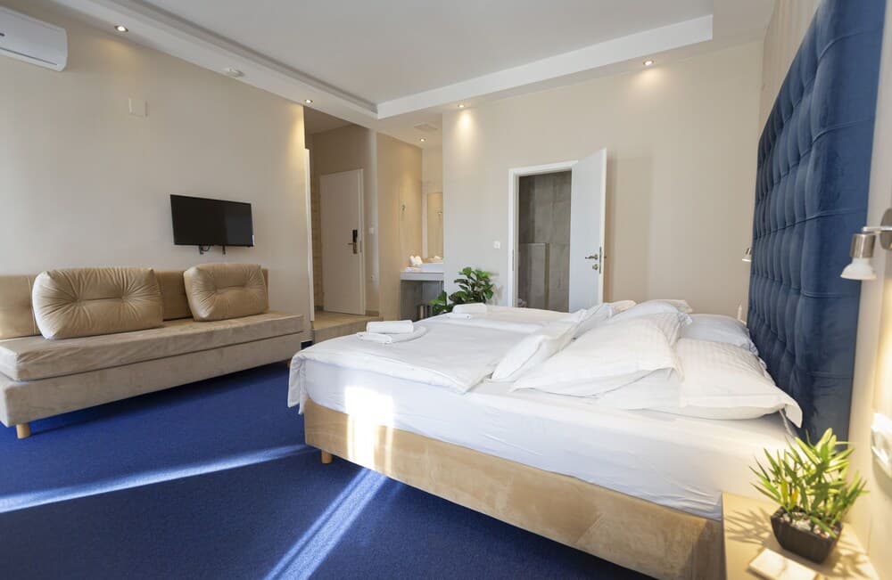 A cozy hotel room features a plush bed with a navy blue headboard, a beige sofa, a flat-screen TV, and soft lighting, creating a serene atmosphere ideal for relaxation and leisure travel.