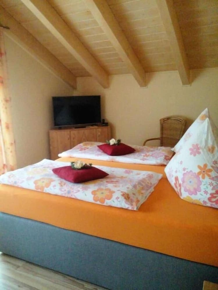 A cozy attic bedroom with wooden beams, vibrant orange bedding, floral patterns, and a flat-screen TV offers a warm and inviting retreat for travelers seeking a charming accommodation experience.