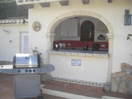 A cozy outdoor kitchen with a tiled counter, built-in barbecue grill, and Mediterranean-style archway invites travelers to enjoy al fresco dining in a serene, sunlit setting.