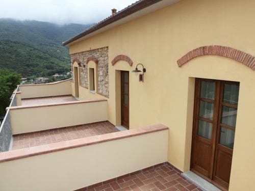A serene balcony of a rustic yellow villa overlooks lush green hills under cloudy skies, featuring arched brick details and wooden doors, ideal for travelers seeking a peaceful retreat in a countryside setting.