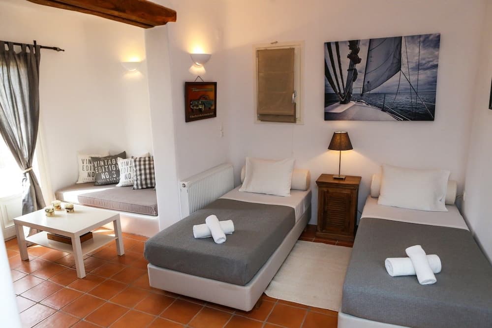 A cozy Mediterranean-style guest room features twin beds with neatly rolled towels, a cushioned bench by the window, warm terracotta flooring, and nautical artwork, ideal for travelers seeking a serene and inviting accommodation.