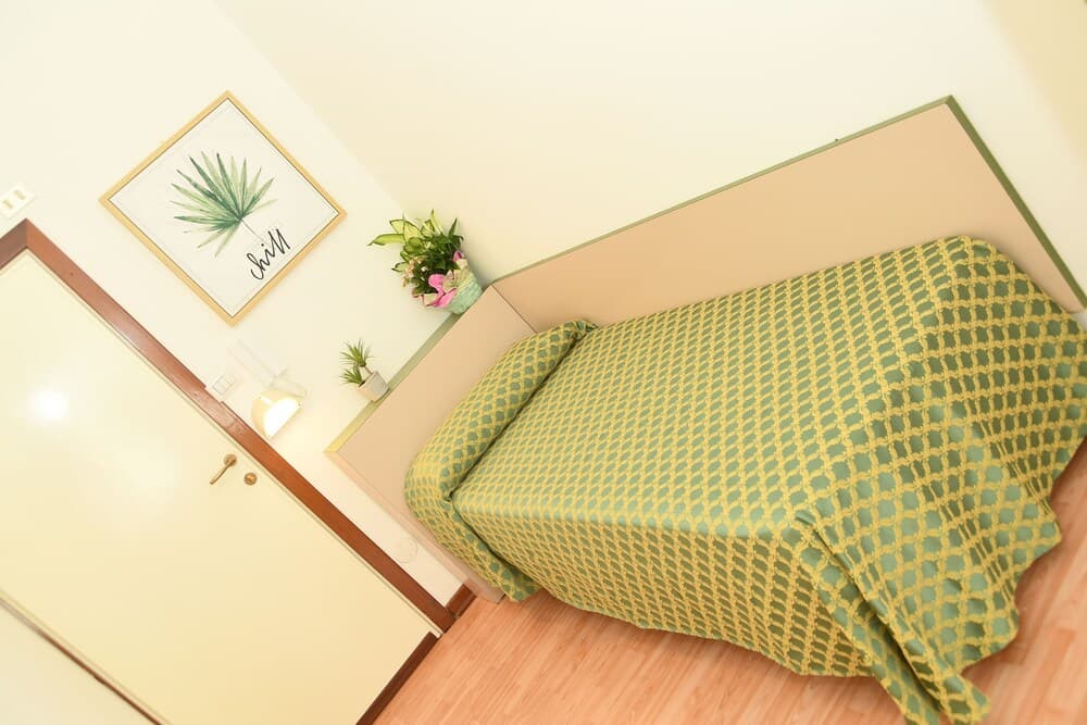 A cozy bedroom with a neatly made bed featuring a green patterned duvet, framed palm leaf art, and a small vase of flowers, ideal for travelers seeking a tranquil and inviting accommodation.