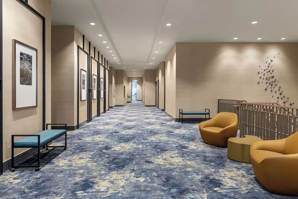 A serene hotel corridor features a calming blue carpet, modern art frames, cozy seating areas, and a butterfly wall sculpture, ideal for travelers seeking comfort and style during their stay.