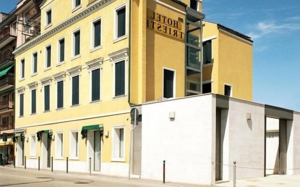 A sunny urban street showcases a vibrant yellow hotel building with classic architecture, green awnings, and modern annexes, ideal for travelers seeking a blend of historic charm and contemporary comfort in a bustling city setting.
