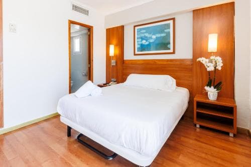 A cozy hotel room features a neatly made bed, wooden furnishings, soft lighting, and a serene painting, creating a welcoming atmosphere ideal for travelers seeking comfort and relaxation.