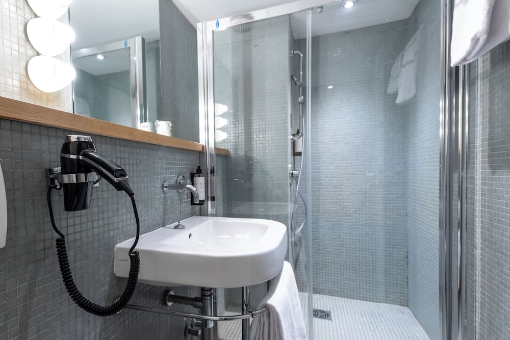 A modern hotel bathroom features sleek glass shower doors, mosaic tiles, a mounted hairdryer, and a bright vanity mirror, ideal for travelers seeking comfort and convenience.