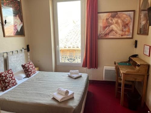 A cozy hotel room with a neatly made bed, vibrant red carpet, wooden desk, and large window offering a view of tiled rooftops, ideal for travelers seeking comfort and charm in a quaint setting.