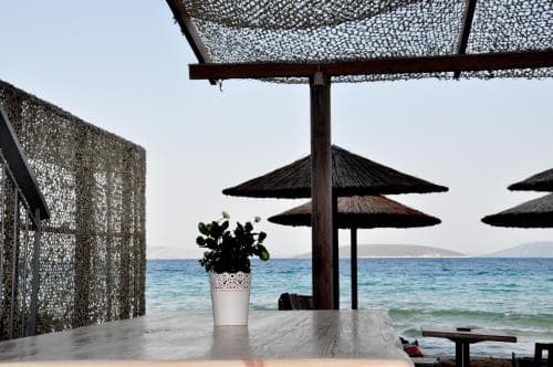 A serene beachside café with wooden tables, thatched umbrellas, and a netted canopy overlooks the tranquil turquoise sea, perfect for a relaxing coastal getaway.