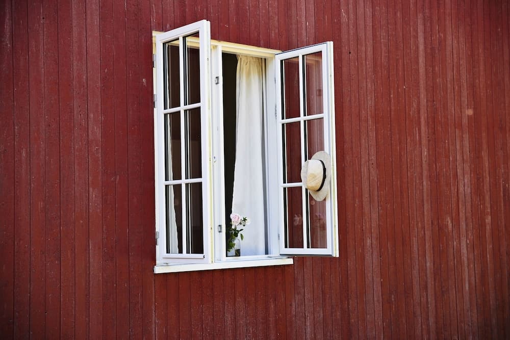 A rustic red wooden house features an open white-framed window adorned with a straw hat and a vase of flowers, evoking a serene countryside charm ideal for travel photography and rural retreats.