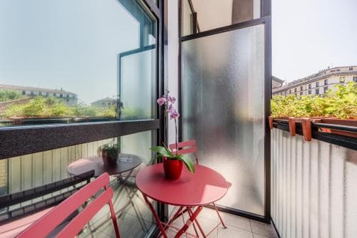 A cozy urban balcony features vibrant red furniture and lush greenery, creating an inviting atmosphere perfect for city travelers seeking a serene retreat.