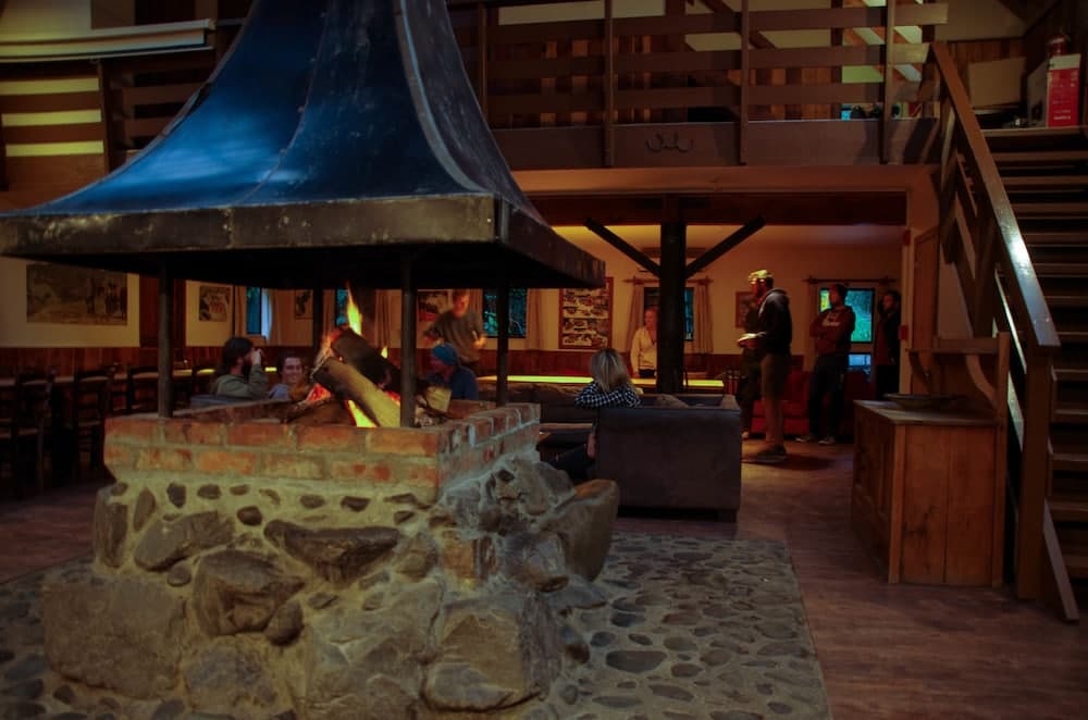 A cozy lodge interior features a central stone fireplace with a blue canopy, surrounded by guests enjoying warm conversations, ideal for winter retreats and family gatherings.
