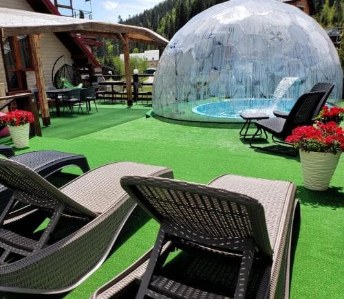 A serene outdoor patio features lounge chairs and a transparent geodesic dome over a hot tub, surrounded by vibrant flowers and artificial turf, ideal for a relaxing mountain retreat.