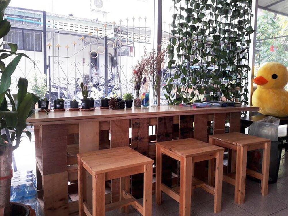 A cozy café corner features rustic wooden tables and stools surrounded by lush greenery and quirky decor like a giant yellow duck, perfect for travelers seeking a relaxed spot to unwind and enjoy a meal.