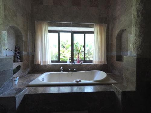 A luxurious bathroom with a large bathtub framed by marble walls and a window offering a serene view of lush greenery, ideal for travelers seeking relaxation and comfort in a tropical retreat.