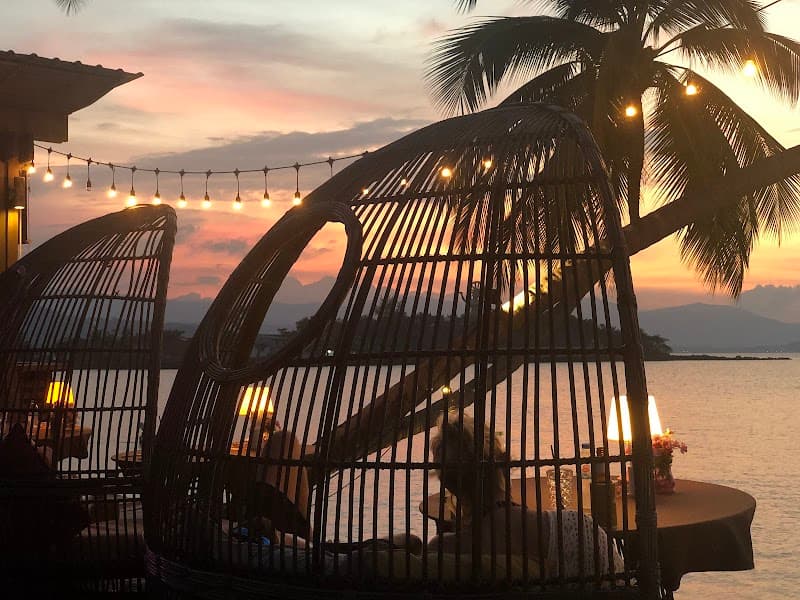 A serene beachside evening at Tango Luxe on Koh Samui, Thailand, features cozy wicker loungers under string lights with a breathtaking sunset backdrop, perfect for a romantic getaway or luxurious relaxation.