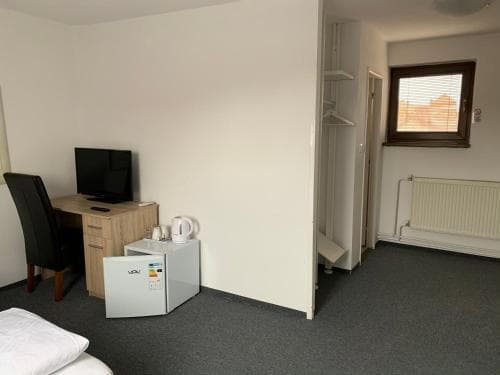 A cozy hotel room features a simple desk setup with a TV, a mini fridge, and a kettle, set against neutral walls and a small window, ideal for travelers seeking a functional and quiet stay.