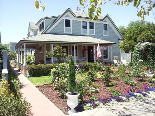 A charming, light blue Victorian-style house with a lush garden and inviting porch offers a serene retreat, perfect for travelers seeking a cozy bed and breakfast experience in a picturesque suburban setting.