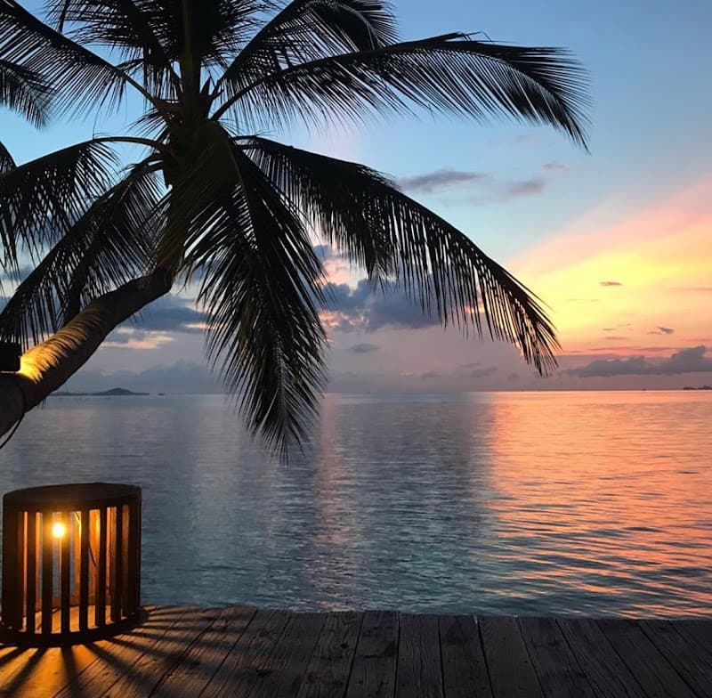 A serene sunset at a tropical beachfront property features silhouetted palm trees and a glowing lantern on a wooden deck, ideal for romantic getaways or tranquil vacations in Thailand.